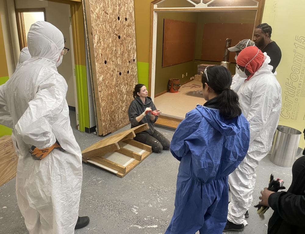 An instructor sits on the floor for a demonstration in front of trainees, who are in personal protective equipment.
